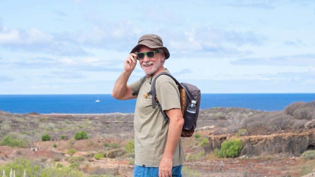 An older, bearded man wearing a hat and backpack smiles in a coastal landscape.