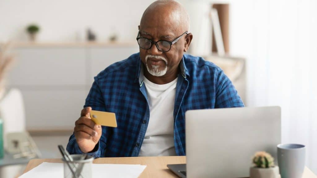 A man with glasses and a beard holds a gold credit card while looking at a laptop.