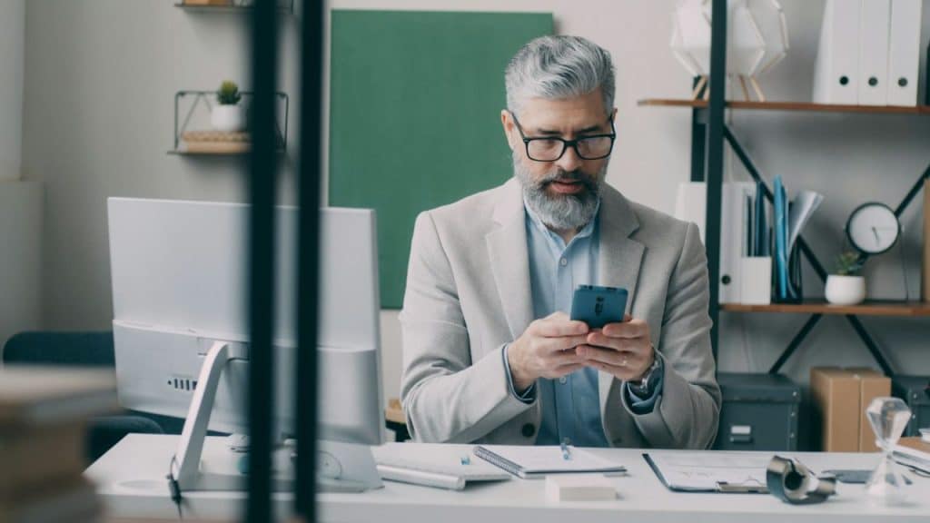 A bearded man in a suit and glasses is looking at his smartphone at an office desk.