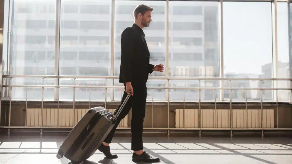 A sharply dressed man walks through an airport terminal, pulling a rolling suitcase.