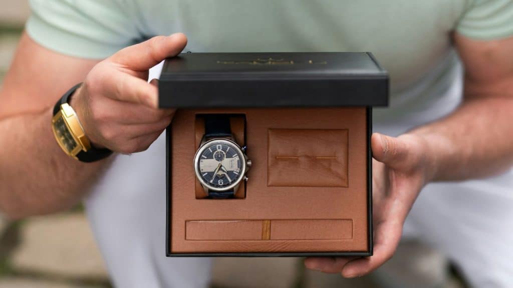 A person's hands open a black box to reveal a blue leather-strapped wristwatch inside.