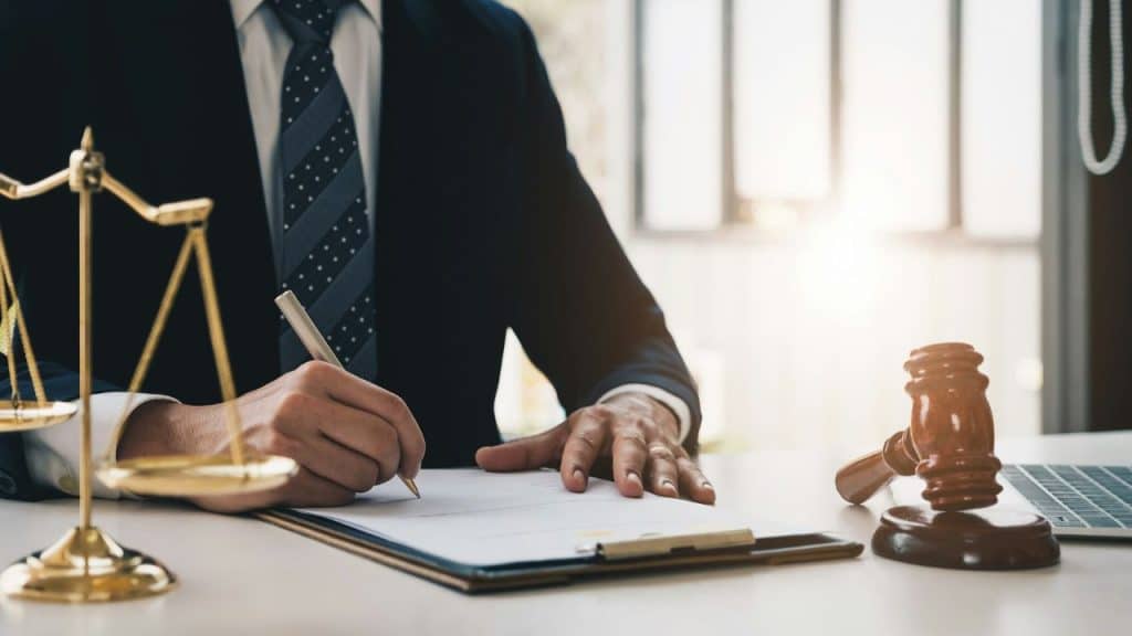 A person in a suit signs a legal document with a gavel and scales of justice nearby.