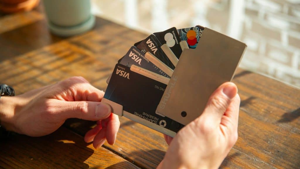 Hands are fanning out multiple credit cards from a slim, metal wallet on a wooden table.