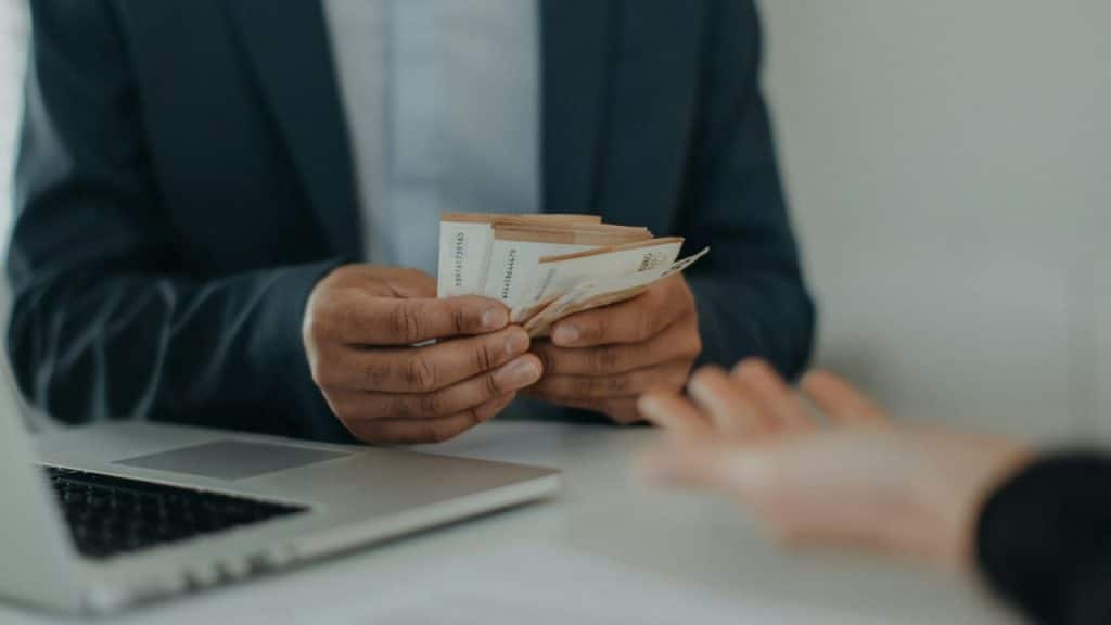 A person in a suit is counting a stack of Euro banknotes while handing them to another person.