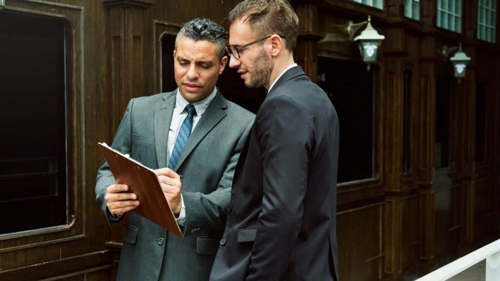 Two businessmen in suits look intently at a clipboard with papers outside a building entrance.