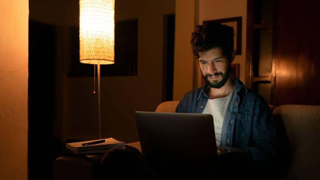 A bearded man is intently using a laptop on a couch at night with a warm lamp.