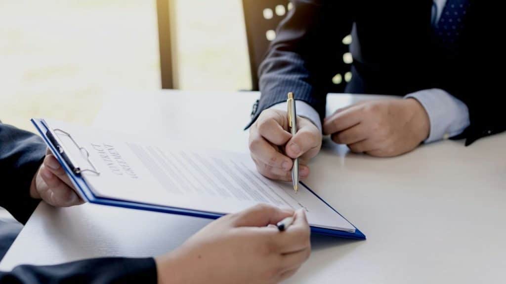 A businessman is signing a contract held by another person across a desk.