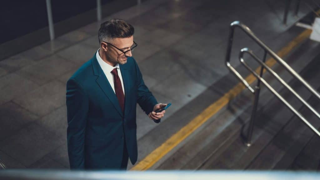 A man in a blue suit and glasses looking at his phone on a staircase.