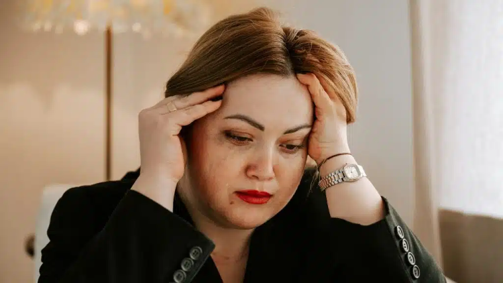A distressed woman with red lipstick holds her head with both hands.