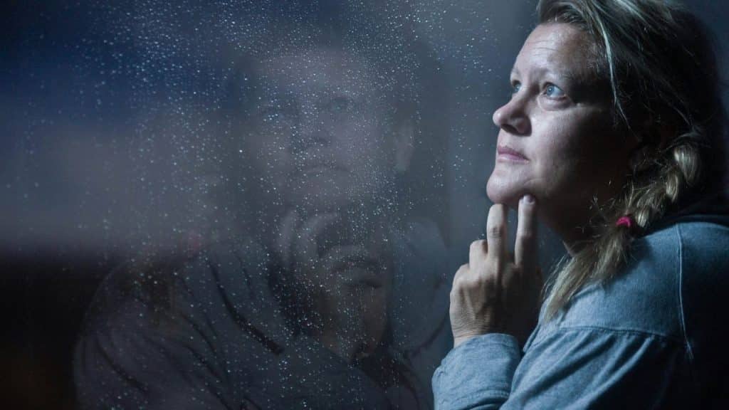 A blonde woman with a pensive expression looks out a rain-streaked window.