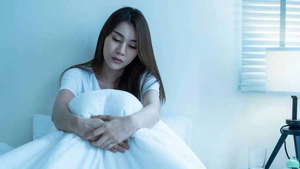 A pensive woman sits up in bed, hugging a white comforter in a dimly lit room.