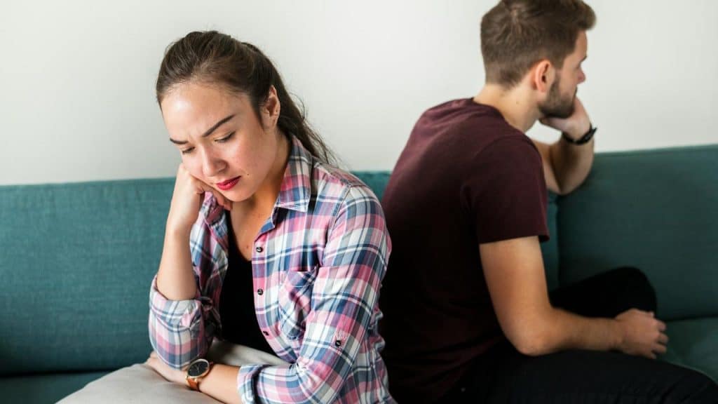 A couple sitting back-to-back on a couch looking upset.