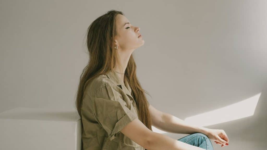 A young woman with long brown hair sits with her eyes closed, looking up.