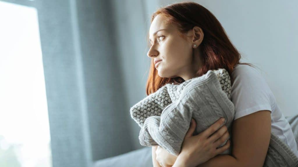 A pensive redheaded woman holds a gray knitted blanket and looks out a window.