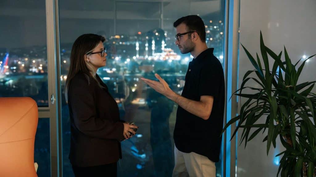 A serious man and woman in glasses are talking next to a large window overlooking a city at night.