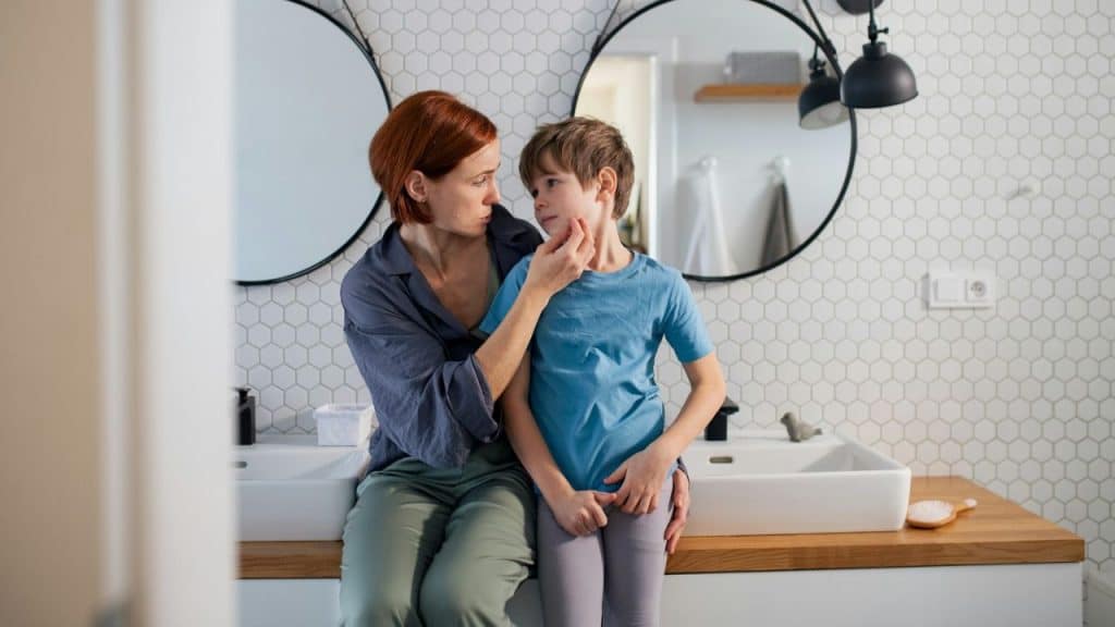 A woman with red hair comforts a young child in a bright, modern bathroom.