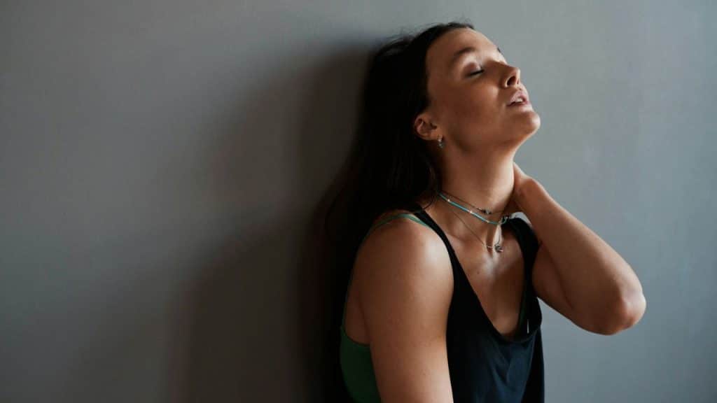 A woman leans against a gray wall, holding her neck with her eyes closed.