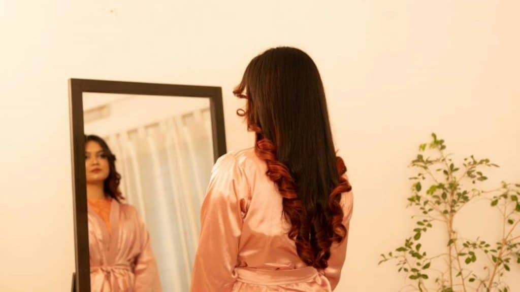 A woman in a silk robe looks at her reflection in a standing mirror.