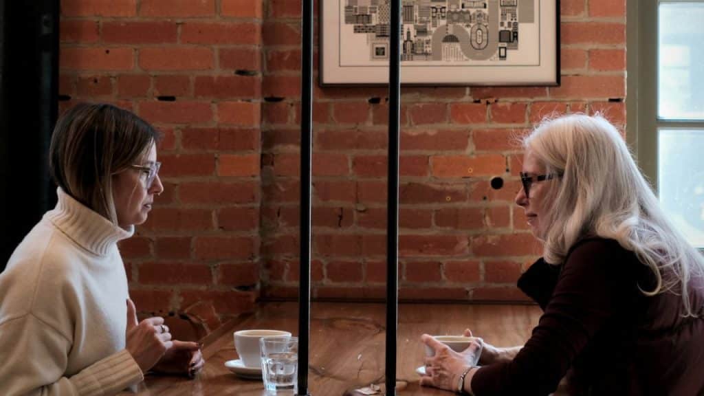 Two women with glasses talk seriously across a table with coffee cups.