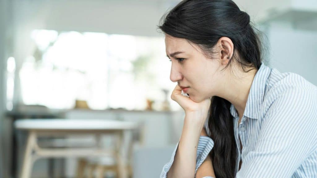 A distraught woman with dark hair rests her chin on her hand, looking down.