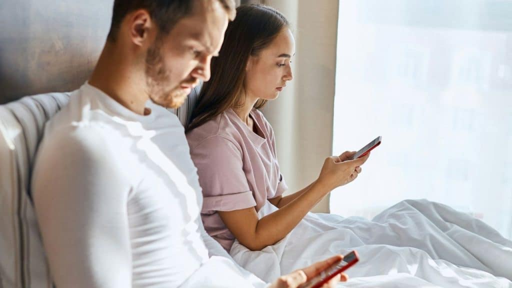 A man and woman sit in bed, both looking intently at their own smartphones.