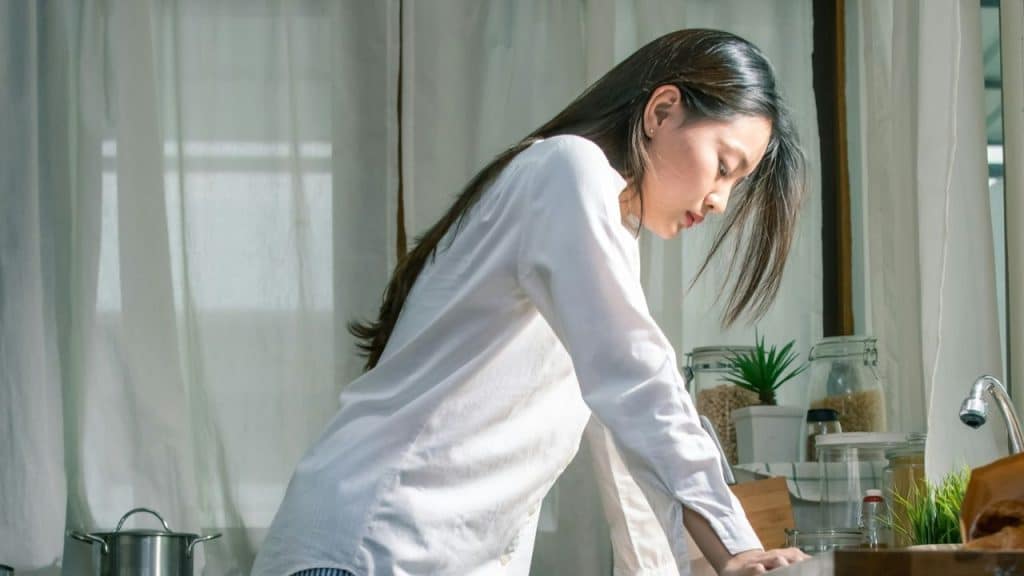 A woman with long dark hair stands leaning on a counter, looking down sadly.