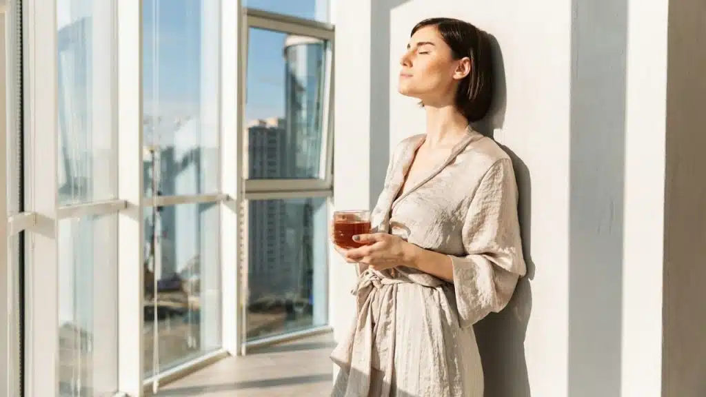 A woman in a silk robe stands by a large window, eyes closed, holding a glass of tea.