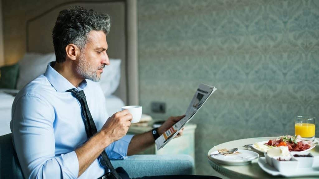 A man in a shirt and tie drinks coffee and reads a newspaper at a breakfast table.