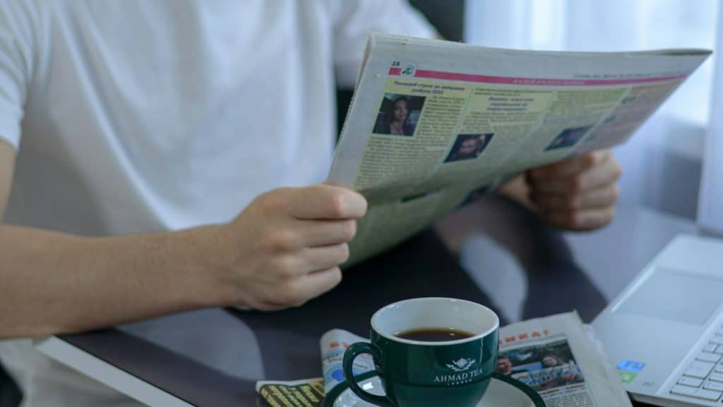 A person in a white shirt is reading a physical newspaper while drinking a cup of tea.