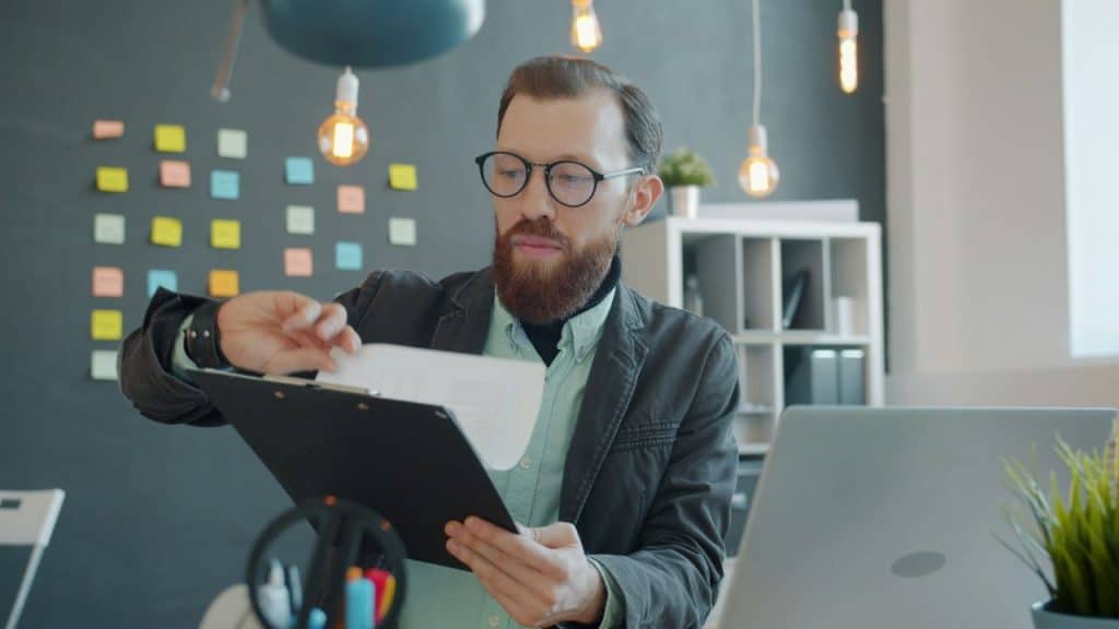 A bearded man in glasses and a blazer looking at documents on a clipboard in an office with industrial lighting.