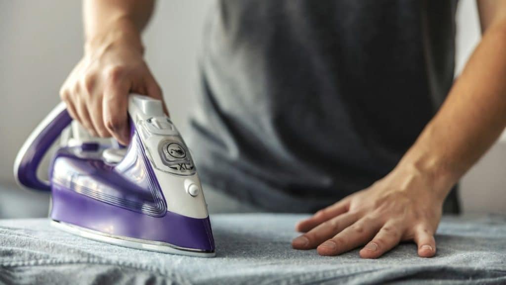 Close-up of a person's hands ironing a gray garment with a purple and white iron.