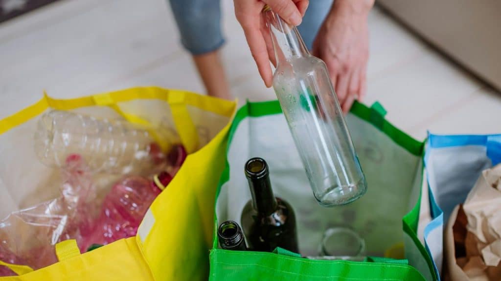 A person sorting recycling by placing a glass bottle into a green reusable bag.