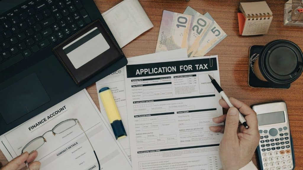 A person filling out a tax application form on a desk covered with paperwork, Canadian cash, and a calculator.