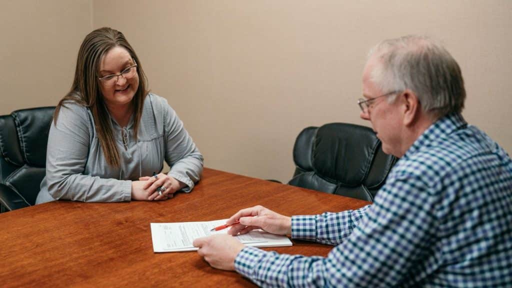 A smiling woman and an older man with glasses sit across a table discussing paperwork in a meeting.