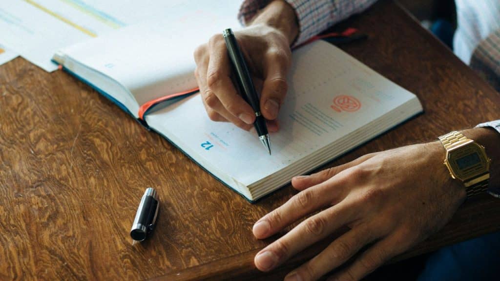 A close-up of a person's hands writing in a planner or diary with a black pen.