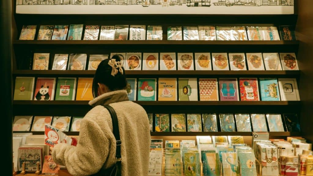 A person wearing a fuzzy jacket looks at a wall display filled with greeting cards in a store.