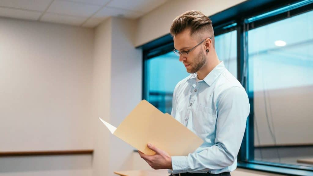 A man with glasses and a beard stands indoors and looks down at a manila folder.