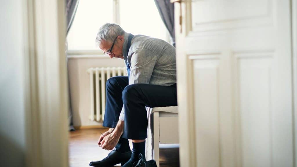 An older man sitting on a bed tying his shoes.