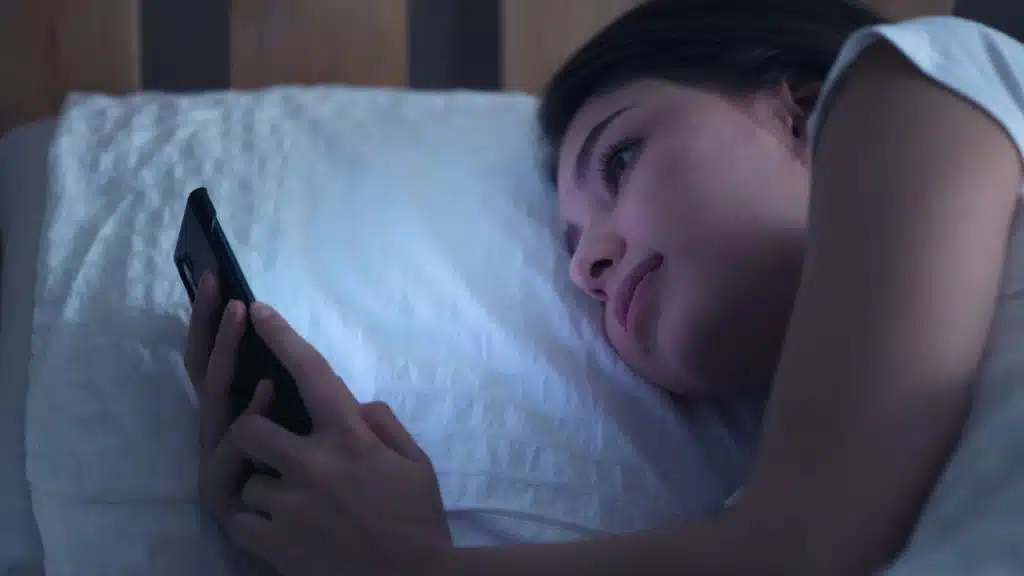 A woman lies in bed in the dark, looking intently at her smartphone.