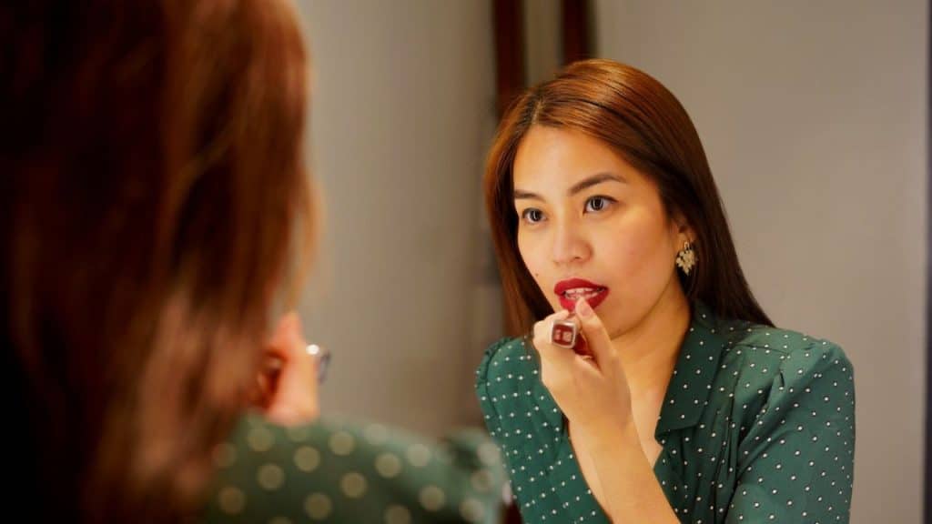 A woman in a green polka-dot top is applying red lipstick in a mirror.