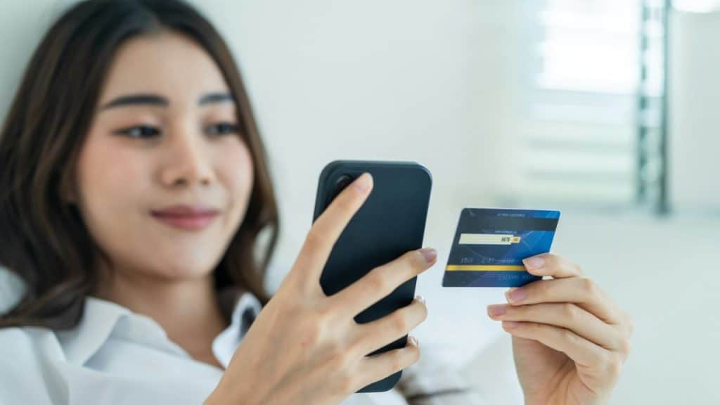 A young woman is holding a smartphone and a blue credit card, looking at the screen.