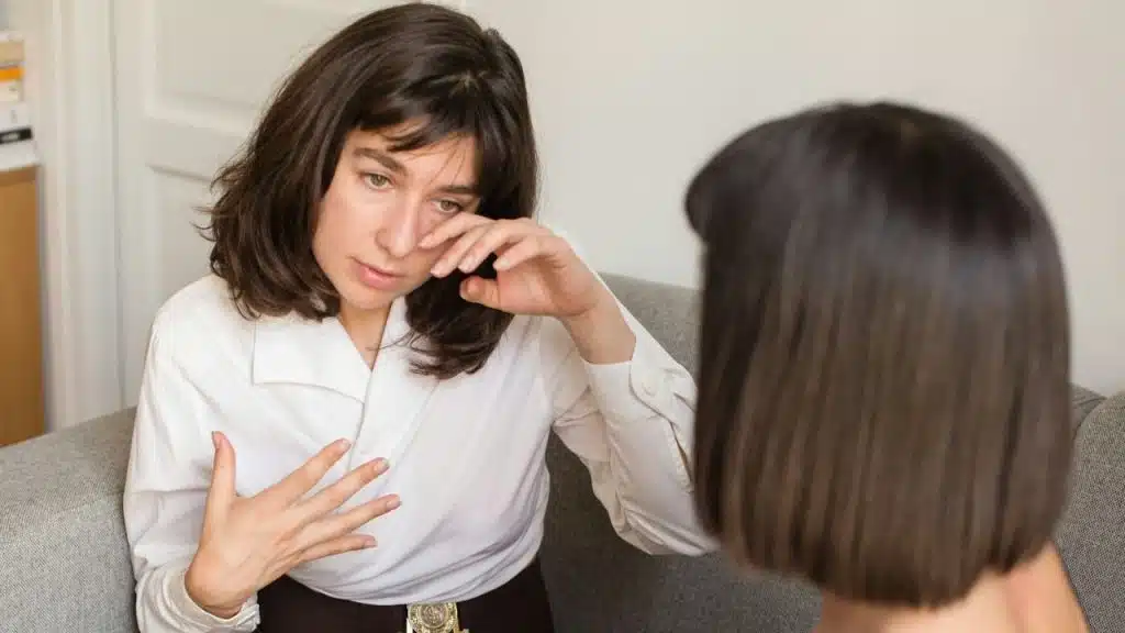 A woman in a white shirt is talking to someone, wiping her eye with one hand.