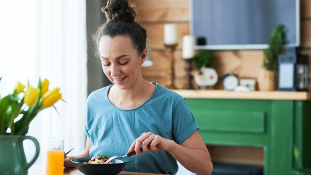 A smiling woman in a blue shirt is eating a meal at a wooden table.