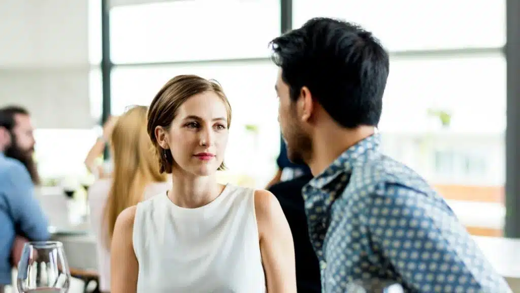 A woman in a white top looks at a man talking to her in a bright room.