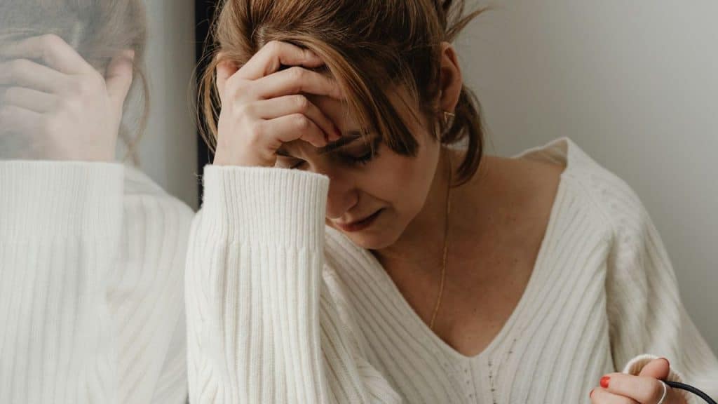 A woman in a white sweater holds her forehead with one hand, looking down in distress.
