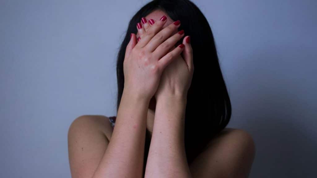 A woman with long dark hair is covering her face with her hands, showing red nail polish.