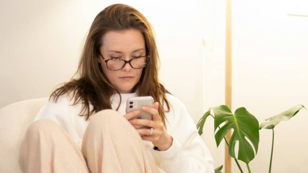 A woman wearing glasses and a white sweater is looking at her smartphone.