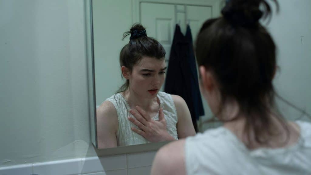 A woman with a bun looks at her reflection, hand on her chest, with an open mouth.