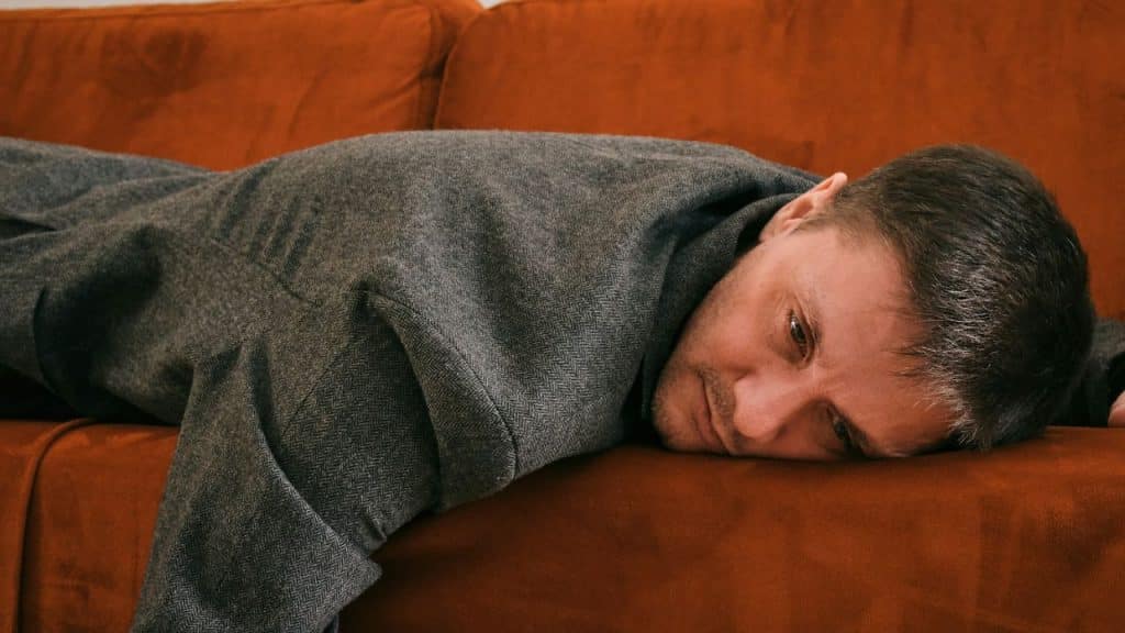 A sad or tired man in a grey jacket is lying face down on an orange couch.