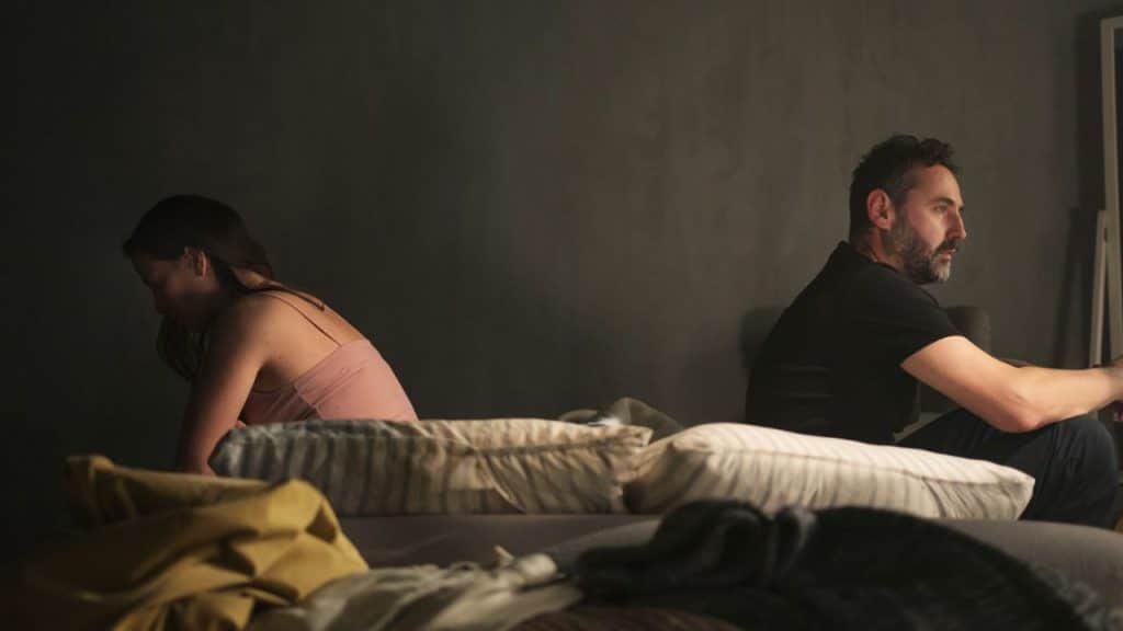A man and woman sit back to back on a bed in a dimly lit room, looking disconnected.